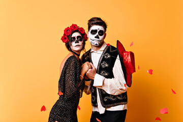 Dead mexican man with sombrero holding girlfriend's hands. Studio shot of romantic couple of zombies isolated on yellow background.
