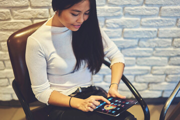 Fototapeta na wymiar Smiling pretty writer sitting on chair and watching funny photos in social networks using modern touch pad enjoying rest indoors. Stylish hipster girl blogging and chatting online in coffee shop