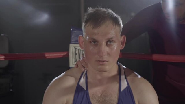 Trainer endorsing young perspiring boxer sitting in corner of boxing ring. Camera approaches to angry face of Caucasian sportsman ready for fight. Combat sport, competition, championship.