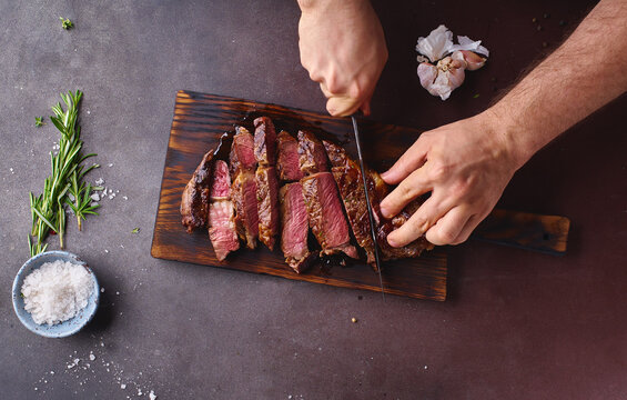 Male hands cutting cooked until medium ribeye steak on wooden cutting board.