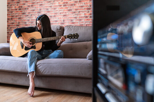 Woman Playing Guitar Sitting On Her Couch At Home And Learning With Online Lessons And Some Masks Are Hanging Due To Containment. Behind It Is A Brick Wall