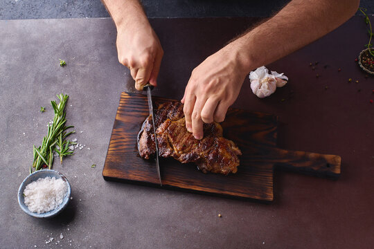 Male hands cutting cooked until medium ribeye steak on wooden cutting board.