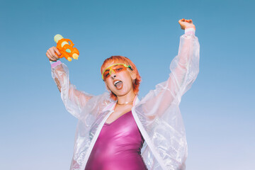 From below excited young woman with water gun in raised arms screaming and celebrating success against cloudless blue sky