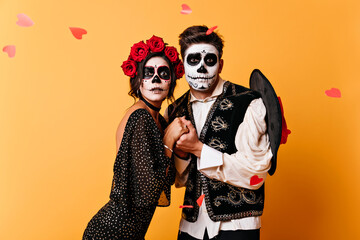 Romantic couple spending valentine's day during zombie photoshoot. Indoor shot of scared young people holding hands on orange background.