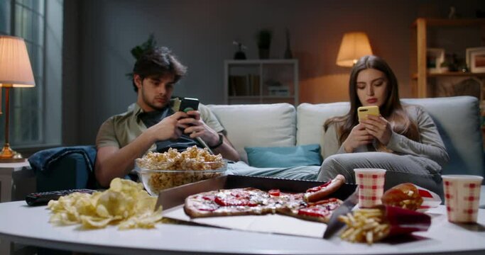Bored young couple is frustrated after hard day, sitting in front of tv but scrolling through social media on their phones and eating unhealthy food 4k footage
