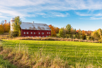 Idyllic rural landscape with an old red wooden barn with a silo under blue sky at sunset. A meadow is in foreground and colourful autumn trees are in background. Countryside of Vermont, United States. © alpegor