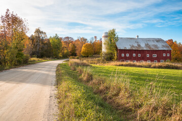 Red traditional barn with silo along an unpaved back road in the countryside of Vermont at sunset. Beautiful autumn colours.