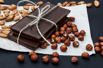 Black chocolate on a black background close-up. Hazelnuts and almonds are scattered in the background. Free space in the background.