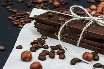 Black chocolate on a black background close-up. Hazelnuts and coffe beans are scattered in the background. Free space in the background. Image of Flat lay. World Chocolate Day.
