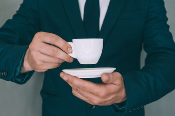 Businessman in a jacket drinks and holds in his hand a cup with a coffee on a gray background.