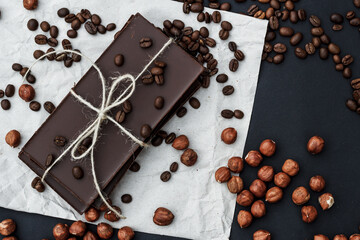 Black chocolate on a black background close-up. Hazelnuts and coffe beans are scattered in the background. Free space in the background. Image of Flat lay. World Chocolate Day.