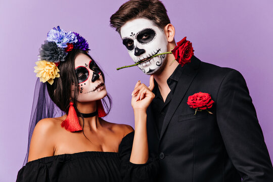Zombie girl looking at her boyfriend with love. Caucasian man with creepy halloween makeup holding rose in his mouth.