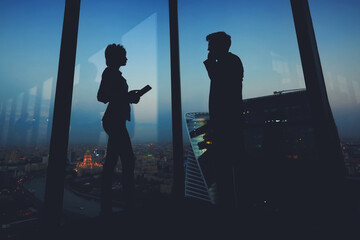 Silhouette of a two young business people standing in modern interior near big window, man entrepreneur having mobile phone conversation while his woman partner using digital tablet during work break