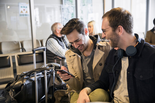 Happy Gay Male Couple With Smart Phone In Airport Departure Area