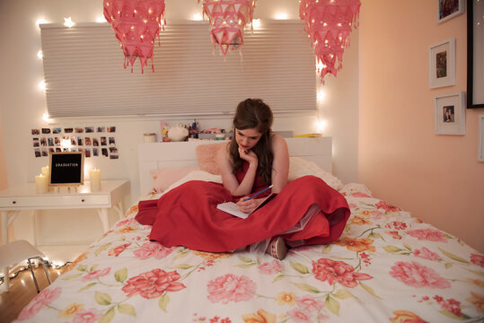 Teenage girl in red dress journaling on bed by graduation sign