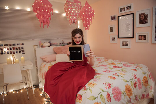 Teenage girl holding graduation sign and smart phone in bedroom