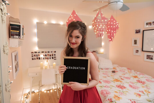 Portrait happy teenage girl holding graduation sign in bedroom