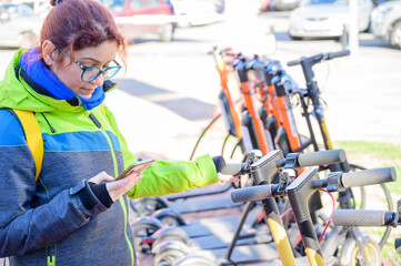 The girl in the jacket uses the mobile application to pay and unlock the rental electric scooter. Close-up female hand with a phone. Woman on eco-friendly transport to move around the city.