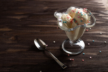 White balls of vanilla ice cream under colorful sprinkles in a dessert bowl. Decorated ice cream with a dessert spoon on a dark wood table.