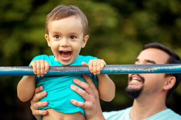 Happy toddler boy doing pull ups exercise, child smiling happily having pulled up on steel bar outdoors in summer day. Cheerful father supporting kid. Success, achievement and healthy family concept