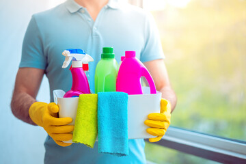 Мan in yellow rubber gloves holding a white plastic box with cleaning products and microfiber rags. Equipment for cleaning and supplies indoors, closeup. Closeup of man househusband