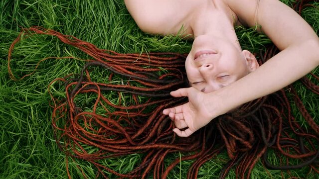 girl with red dreadlocks lying on the green grass, squinting from the sun, smiling. view from above.
