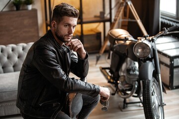 Fototapeta na wymiar Handsome young man in leather jacket looking away while sitting on the stool
