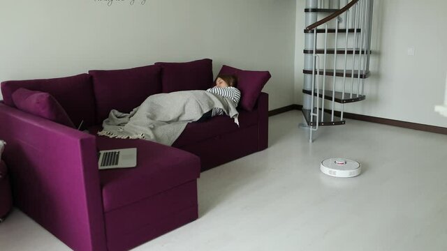 Young woman is resting on sofa while robotic vacuum cleaner is vacuuming apartment. Modern technologies and household concept