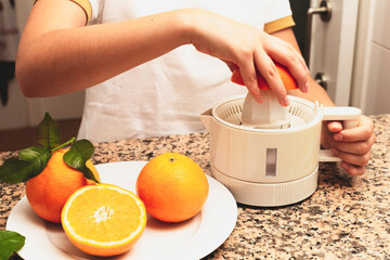 A teenager girl in an apron squeezes juice from oranges using an electric juicer on a table in the kitchen.