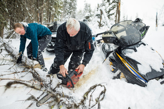 Elderly man and son using chainsaw to clear trails while snowmobiling.