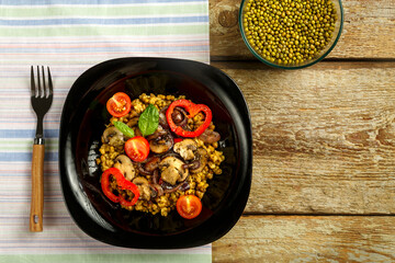 On a wooden table is a black plate of boiled mung bean with vegetables and mushrooms on a napkin and a fork. Copy space.