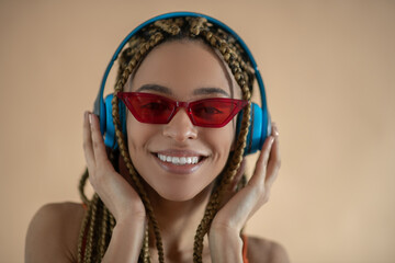 Smiling young African American female in red eyeglasses, touching blue headphones and listening to music