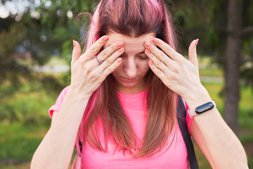 woman have a headache pain outdoors. girl has pink hair and a pink colored shirt. female closed her eyes and holds hands on head.