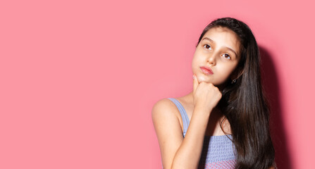 Studio portrait of thoughtful child girl, holding hand on chin, looking up. Background of pastel pink color with copy space.