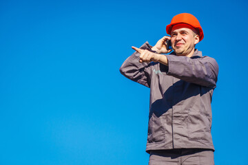 Construction worker in a helmet against the blue sky talking on the phone. Contractor control according to plan. Builder engineer helmet works at construction site. Copy space.