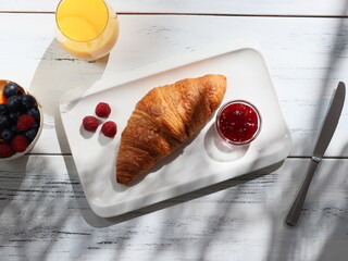 one croissant with berry jam on a white rectangular plate lies on a white wooden table with  and fresh orange juice.