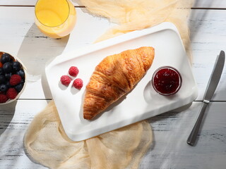 one croissant with berry jam on a white rectangular plate lies on a white wooden table with  and fresh orange juice.