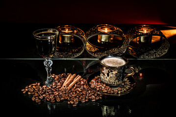 Cup of coffee, with roasted beans and cinnamon sticks around. Glass with black alcoholic drink and behind candle holder with burning candles on the black background. Still life photo. Luxury concept.