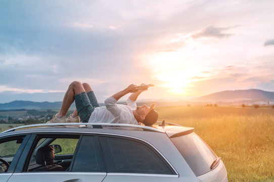 Mature age Man lying on car roof and reading the paper bestseller book. He stopped his auto on high grass meadow with beautiful valley view before the sunset. Reading hobby or education concept image.
