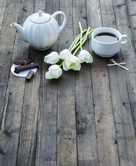 Tea time. Cup of black tea, teapot, chocolate and white tulips flowers on rustic dark brown wooden background. Copy space
