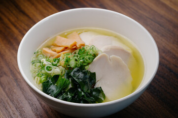 chicken broth ramen noodle soup with poached chicken breast, bamboo shoots, seaweed, scallion in a white bowl on wooden table