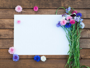 Colorful bouquet of summer garden flowers. Cornflowers with blank paper for greeting message on brown wooden table. Vintage floral background. Floral mock up with multicolored flowers. Copy space
