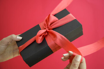 Girl holds a gift box and unties the bow. a girl opens a gift box. Gift box on a red background. Girl unties the tape from the box.
