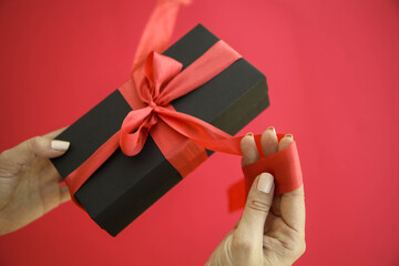 Girl holds a gift box and unties the bow. a girl opens a gift box. Gift box on a red background. Girl unties the tape from the box.
