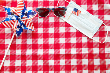 Obraz premium July 4 background. USA flags, sunglasses, and medical mask on red tablecloth. Independence Day of America during coronavirus covid-19 pandemic quarantine. Flat lay, top view, daylight. Copy space text
