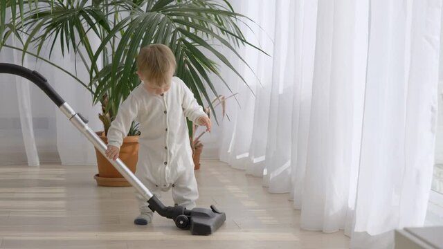 Funny baby child trying hard to learn to use vacuum cleaner, step and falling down