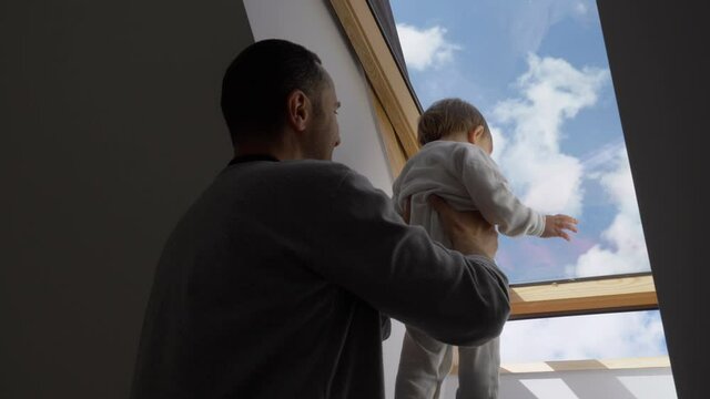 Father hold baby child and look through attic window, blue sky, fresh air, parent rising up child to discover nature, slow motion