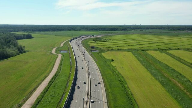 Asphalt ringroad. Speedway with cars through field near forest. Aerial view from copter