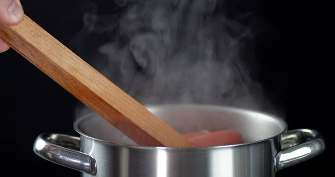 Male hand holding forceps gets boiled sausage from pan.