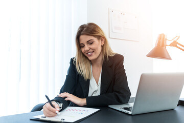 Young confident businesswoman sitting in her bright office while looking at the clipboard on the office table.